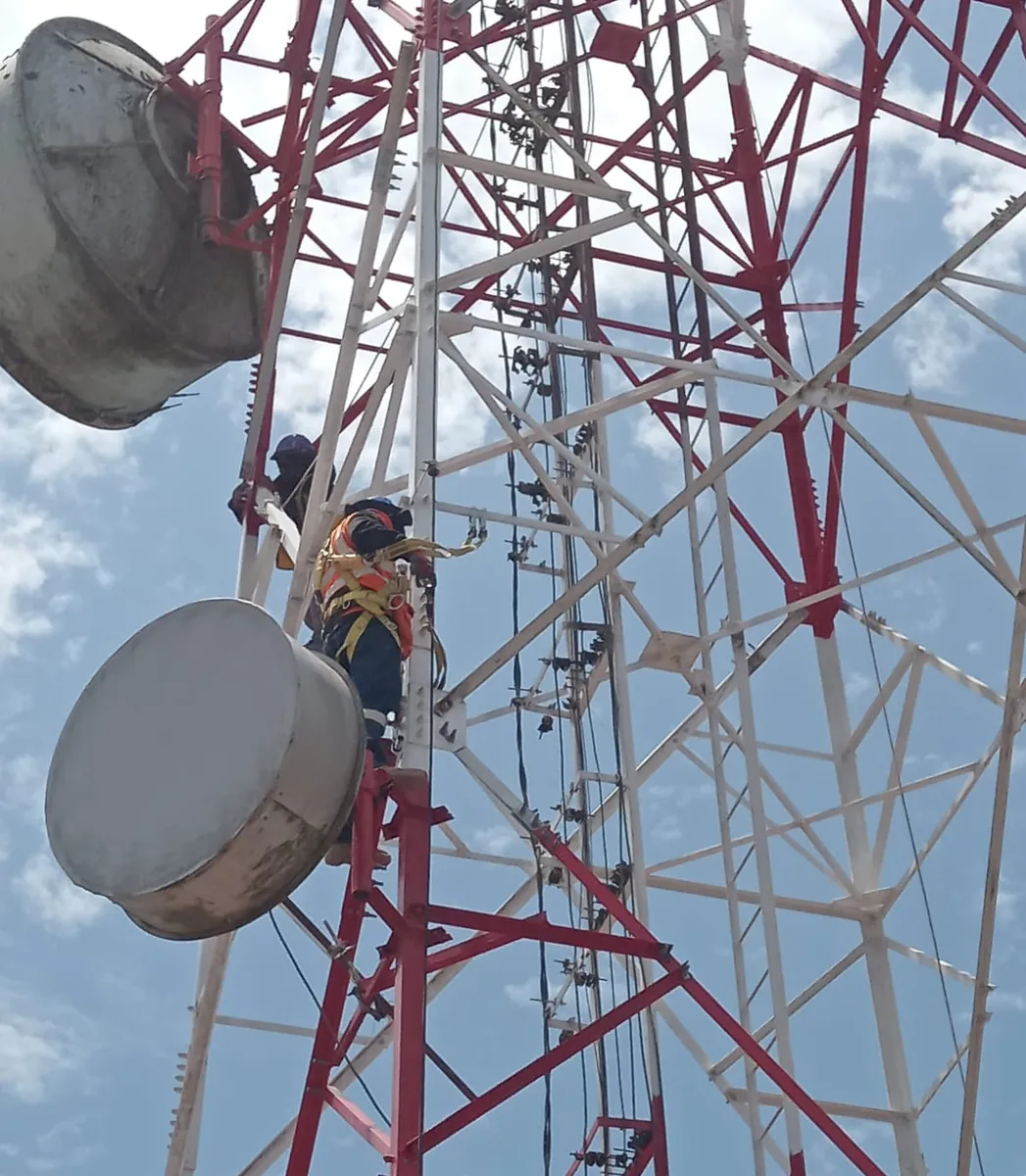 Técnico asegurado con arnés trabajando entre las antenas de una torre de celosía roja y blanca.