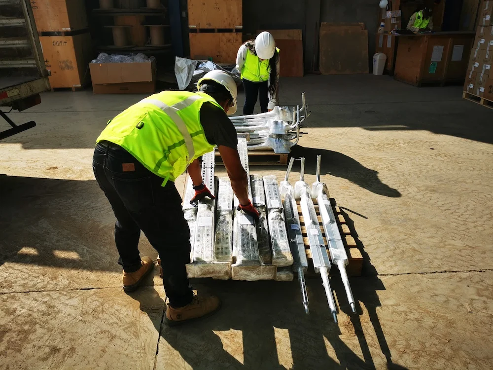 Operario con chaleco reflectante ordenando piezas metálicas galvanizadas en el almacén antes del despacho.